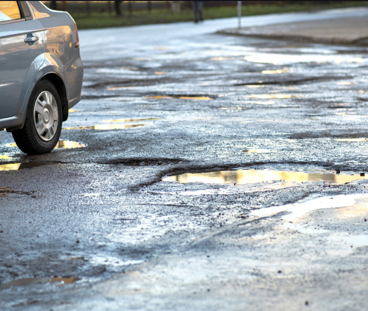 A silver car is parked on the edge of a road riddled with large, water-filled potholes - https://www.american-insurance-center.com/