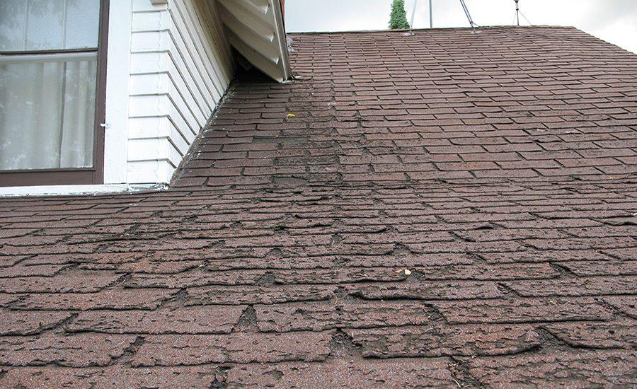 Weathered brown asphalt shingle roof with curling, cracked shingles beside a white-sided dormer window